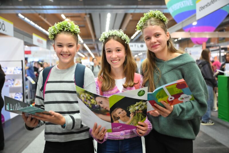 Drei junge Mädchen mit Blumenkränzen am Kopf halten Messebroschüren in den Händen