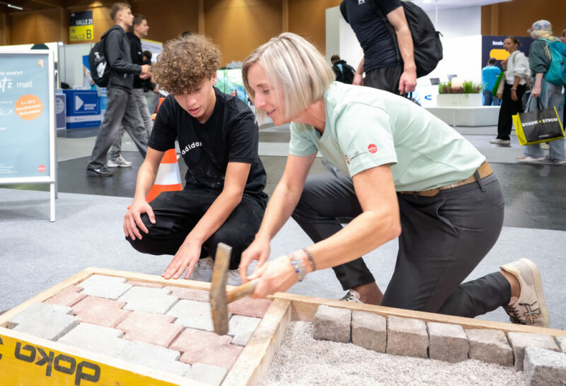 Ausstellerin zeigt Jugendlichem das Legen von Pflastersteinen bei Messestand