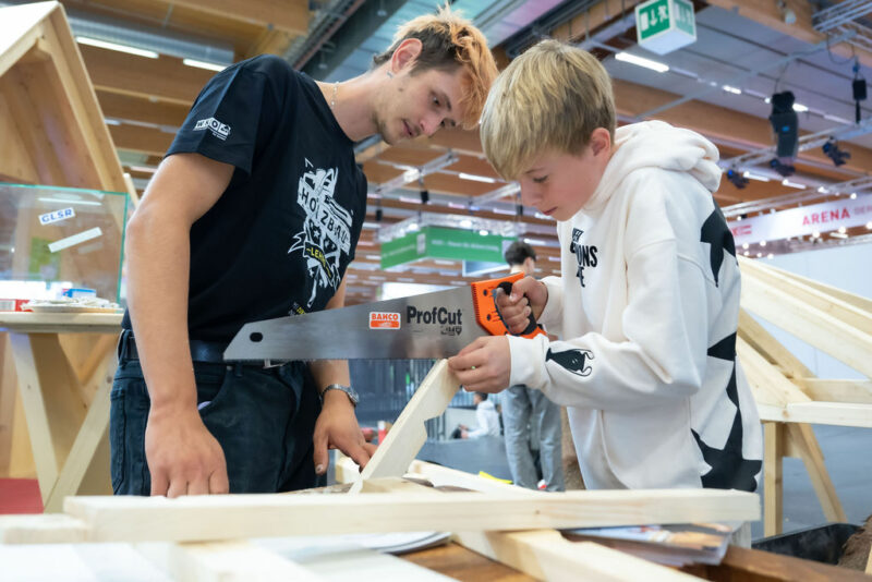 Junge sägt unter Aufsicht eines Ausstellers ein Werkstück bei Messestand
